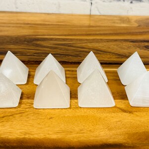 Puede incluir: Ocho pir&aacute;mides de cristal de selenita blanca dispuestas en fila sobre una superficie de madera.
