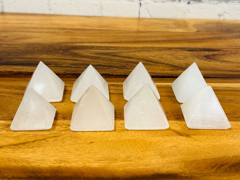 Puede incluir: Ocho pir&aacute;mides de cristal de selenita blanca dispuestas en fila sobre una superficie de madera.