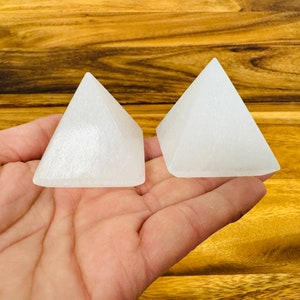 May include: Two white selenite crystal pyramids on a wooden surface. The pyramids are smooth and have a slightly translucent appearance.