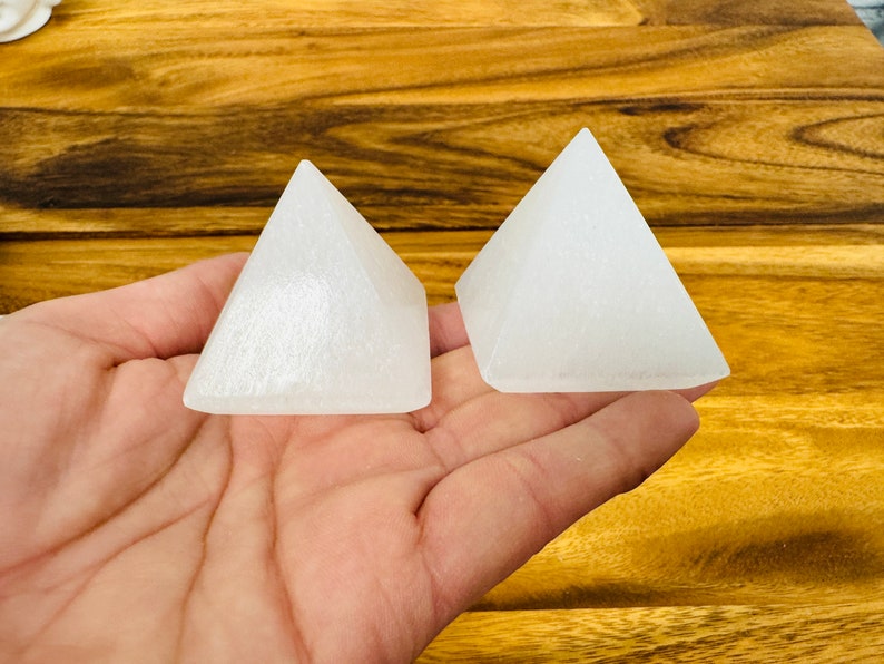 May include: Two white selenite crystal pyramids on a wooden surface. The pyramids are smooth and have a slightly translucent appearance.