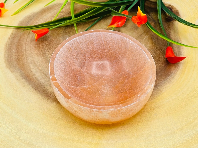 May include: A light pink, round, selenite bowl with a smooth, polished surface. The bowl is sitting on a wooden surface.