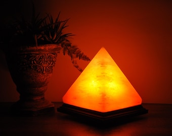 May include: A pyramid-shaped salt lamp emitting a warm orange glow, resting on a dark square base. The lamp features a translucent, crystalline texture with visible internal variations, casting soft light in a dim environment.