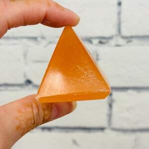 May include: A small, orange, pyramid-shaped crystal. The crystal is smooth and polished, with a slight sheen.