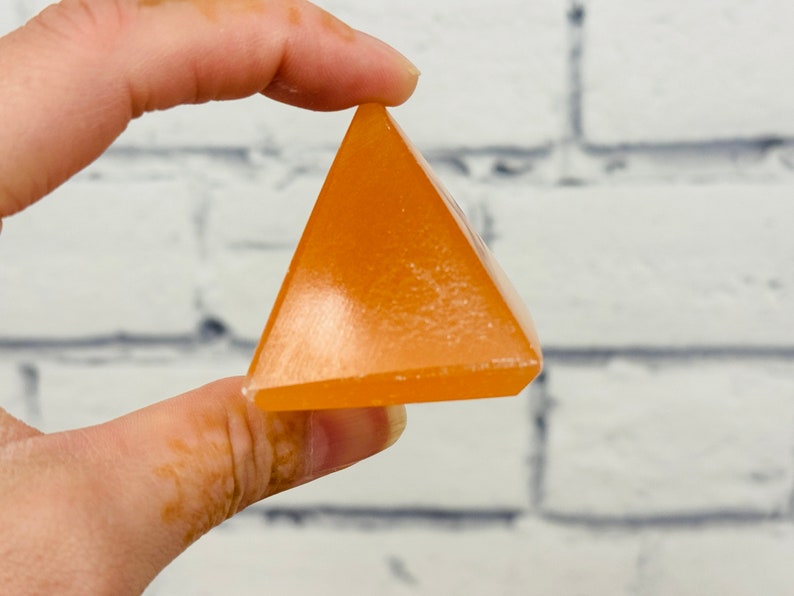 May include: A small, orange, pyramid-shaped crystal. The crystal is smooth and polished, with a slight sheen.