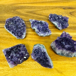 May include: Six amethyst crystal clusters of varying sizes and shapes, displayed on a wooden surface. The crystals are a deep purple color and have a natural, rough texture.