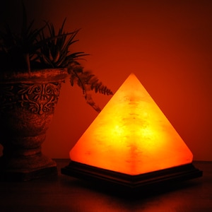 May include: A glowing, pyramid-shaped Himalayan salt lamp on a dark wooden base. The lamp emits a warm, orange light, creating a cozy ambiance. A decorative vase with plants is in the background, adding to the room's decor.