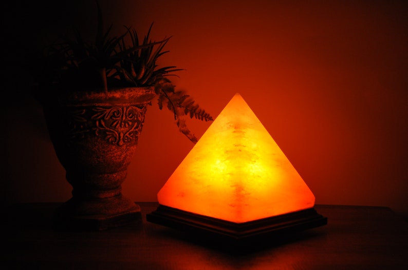 May include: A glowing, pyramid-shaped Himalayan salt lamp on a dark wooden base. The lamp emits a warm, orange light, creating a cozy ambiance. A decorative vase with plants is in the background, adding to the room's decor.