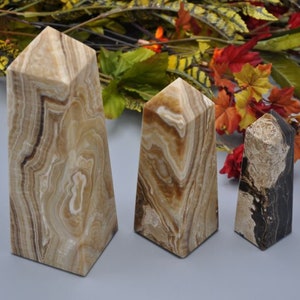 May include: Three stone obelisks with a pyramid top. The obelisks have a beige and brown marbled pattern. The obelisks are different sizes, with the largest being approximately 8 inches tall. The background has fall foliage.