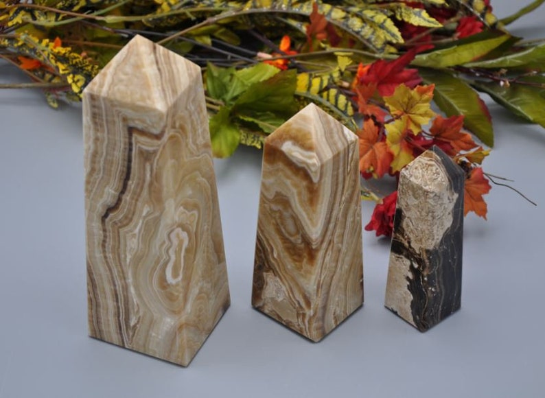 May include: Three stone obelisks with a pyramid top. The obelisks have a beige and brown marbled pattern. The obelisks are different sizes, with the largest being approximately 8 inches tall. The background has fall foliage.