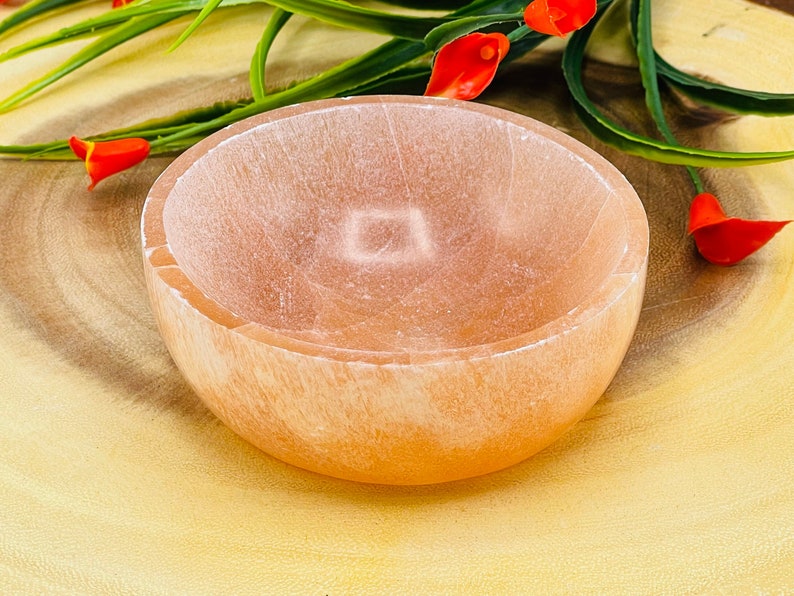May include: A light orange selenite bowl with a smooth, polished surface. The bowl is round and has a slight lip around the edge. It is sitting on a wooden surface.