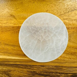 May include: A white selenite charging plate, a circular disc with a textured surface, on a wooden background.