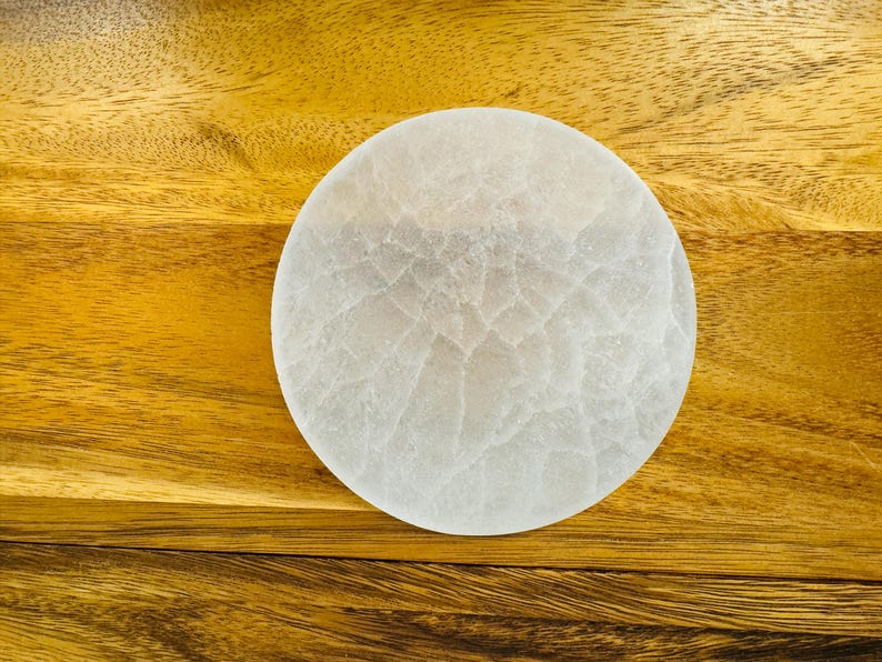 May include: A white selenite charging plate, a circular disc with a textured surface, on a wooden background.