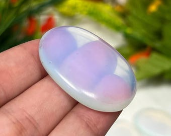 Könnte beinhalten: Ein ovaler, glatter, durchscheinender Cabochon-Stein mit milchig weißem Aussehen und einem sanften, irisierenden rosa und blauen Schimmer, der durch die polierte Oberfläche sichtbar ist. Der Stein hat eine gewölbte Oberseite und eine flache Basis.