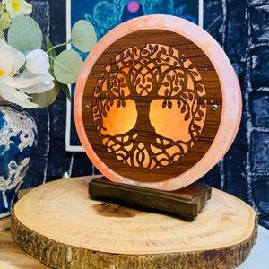 May include: A Himalayan salt lamp with a carved wooden tree of life design. The lamp is lit and glows orange. The lamp is on a wooden base and is sitting on a wooden slice.