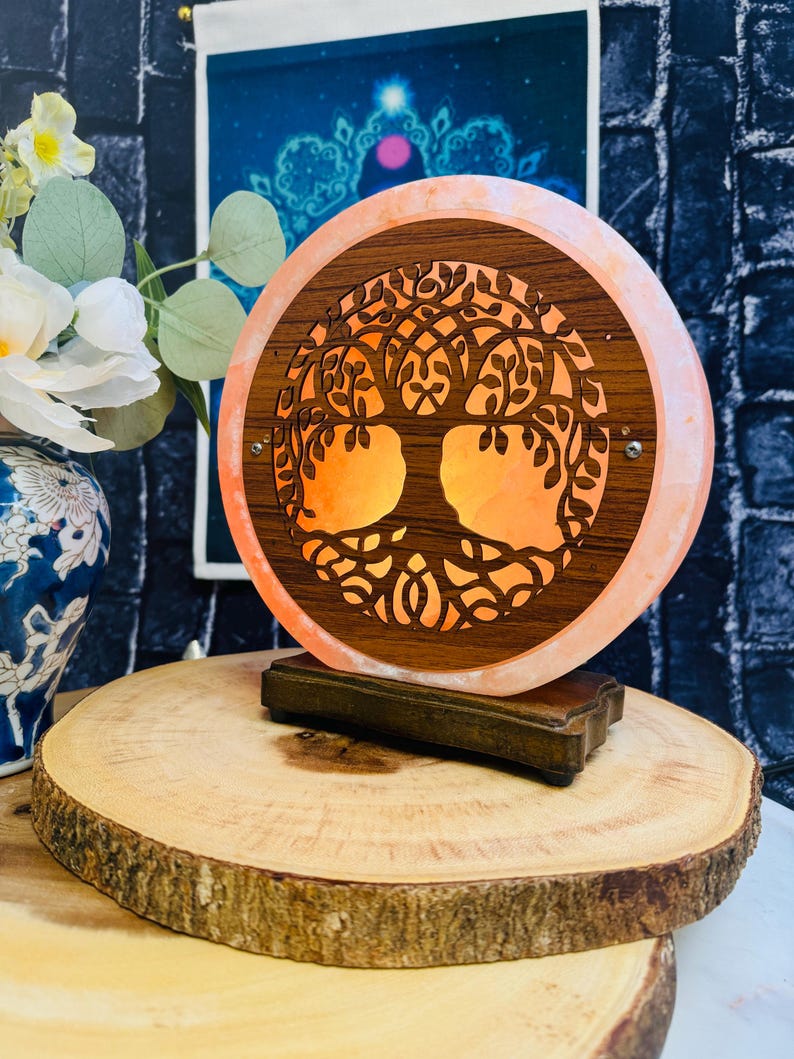 May include: A Himalayan salt lamp with a carved wooden tree of life design. The lamp is lit and glows orange. The lamp is on a wooden base and is sitting on a wooden slice.