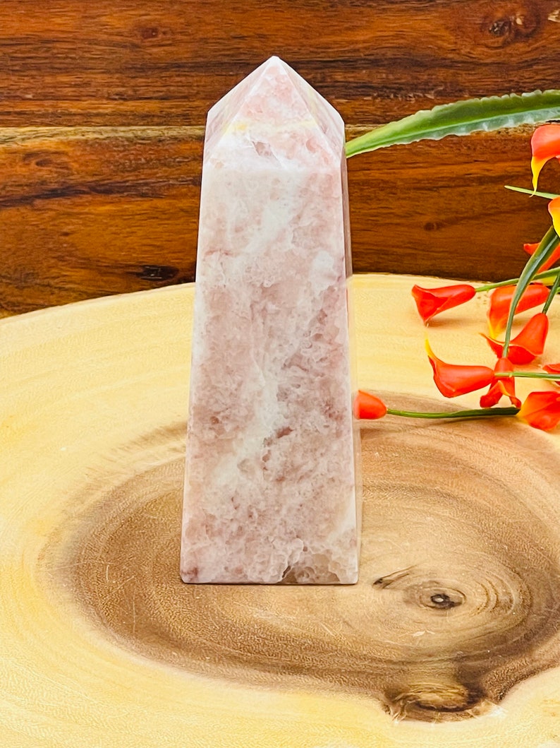Peut inclure: Une pointe de cristal rose et blanche, &eacute;galement appel&eacute;e ob&eacute;lisque de cristal, repose sur une surface en bois. Le cristal a une finition lisse et polie et est en forme de pyramide.