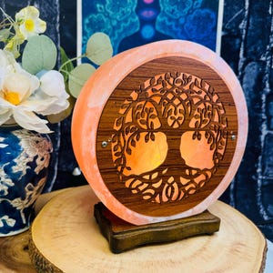 May include: A round Himalayan salt lamp with a wooden tree of life design. The lamp is lit and glows orange. The lamp is sitting on a wooden base.