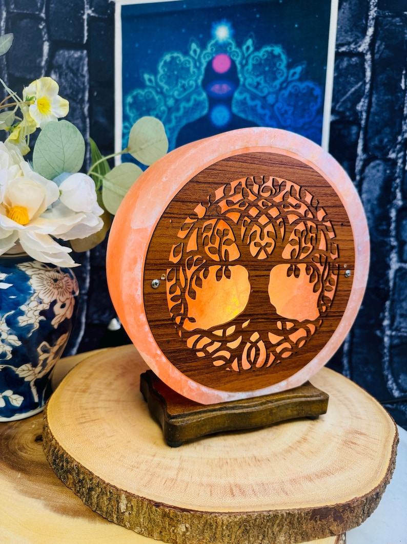 May include: A round Himalayan salt lamp with a wooden tree of life design. The lamp is lit and glows orange. The lamp is sitting on a wooden base.