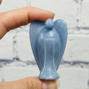 May include: A light blue angel figurine carved from selenite crystal. The angel has a rounded head and wings, and is standing on a flat base.