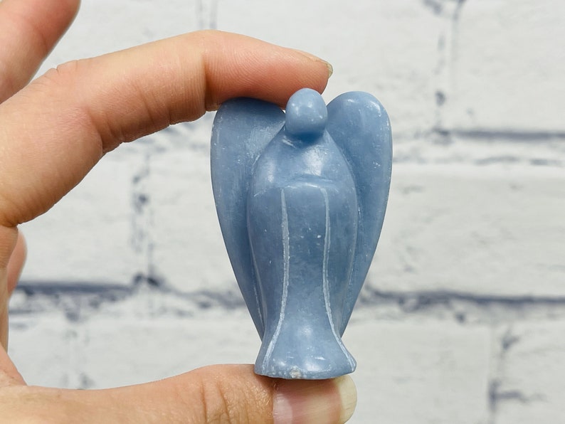 May include: A light blue angel figurine carved from selenite crystal. The angel has a rounded head and wings, and is standing on a flat base.