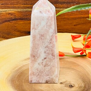 Peut inclure: Une pointe de cristal rose et blanche, &eacute;galement connue sous le nom d'ob&eacute;lisque de cristal, est pr&eacute;sent&eacute;e sur une surface en bois. Le cristal a une finition lisse et polie et une pointe pointue.
