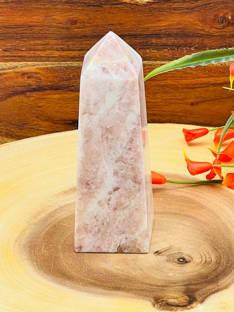 Peut inclure: Une pointe de cristal rose et blanche, &eacute;galement connue sous le nom d'ob&eacute;lisque de cristal, est pr&eacute;sent&eacute;e sur une surface en bois. Le cristal a une finition lisse et polie et une pointe pointue.