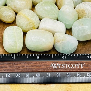 Op de afbeelding: Een verzameling gladde, witte en groene getrommelde stenen, waarschijnlijk celesteen, gerangschikt op een houten oppervlak met een liniaal als schaal. De stenen hebben een diameter van ongeveer 2,5 cm.
