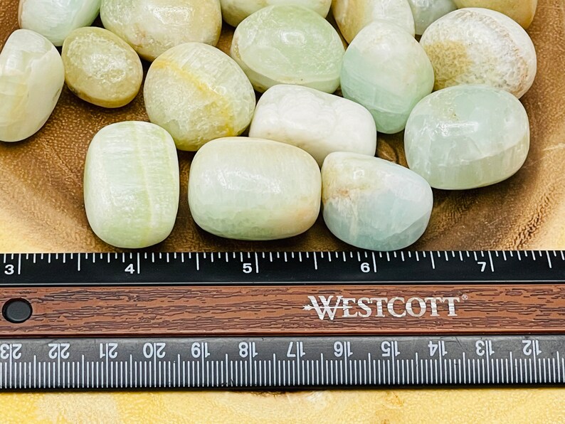 Op de afbeelding: Een verzameling gladde, witte en groene getrommelde stenen, waarschijnlijk celesteen, gerangschikt op een houten oppervlak met een liniaal als schaal. De stenen hebben een diameter van ongeveer 2,5 cm.