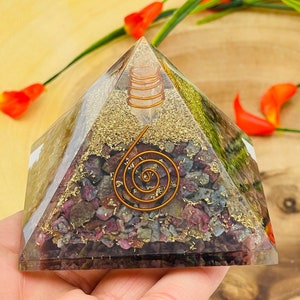 May include: A clear pyramid with a copper wire spiral inside. The pyramid is filled with small pink, green, and gold crystals.