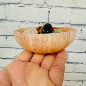 May include: A light pink selenite bowl with a smooth, polished surface. The bowl is filled with a variety of colourful gemstones, including clear quartz, tiger's eye, and amethyst.
