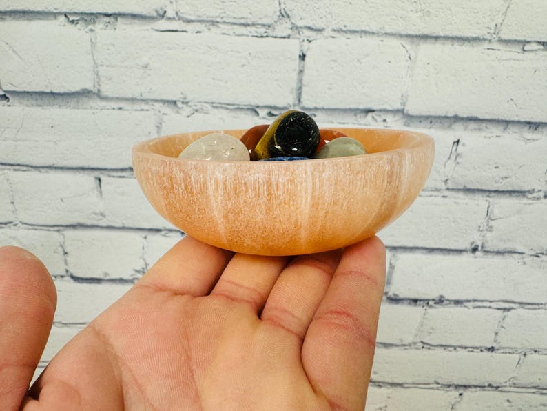 May include: A light pink selenite bowl with a smooth, polished surface. The bowl is filled with a variety of colourful gemstones, including clear quartz, tiger's eye, and amethyst.