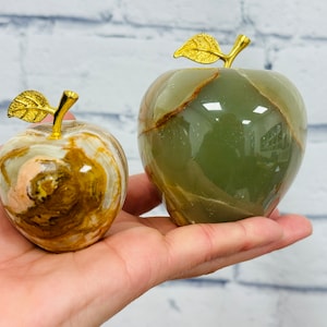 May include: Two decorative apple sculptures. One is a mix of beige, brown, and gold tones, and the other is a deep green with brown veining. Both have gold-colored leaf and stem details, and are held in a hand.