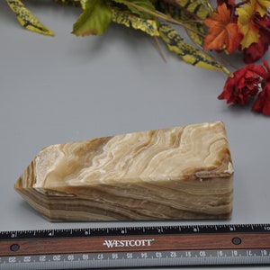 May include: A rectangular, beige and brown onyx crystal with a layered pattern. The crystal is cut with angled sides and a flat top. A ruler with inch measurements is in the foreground.