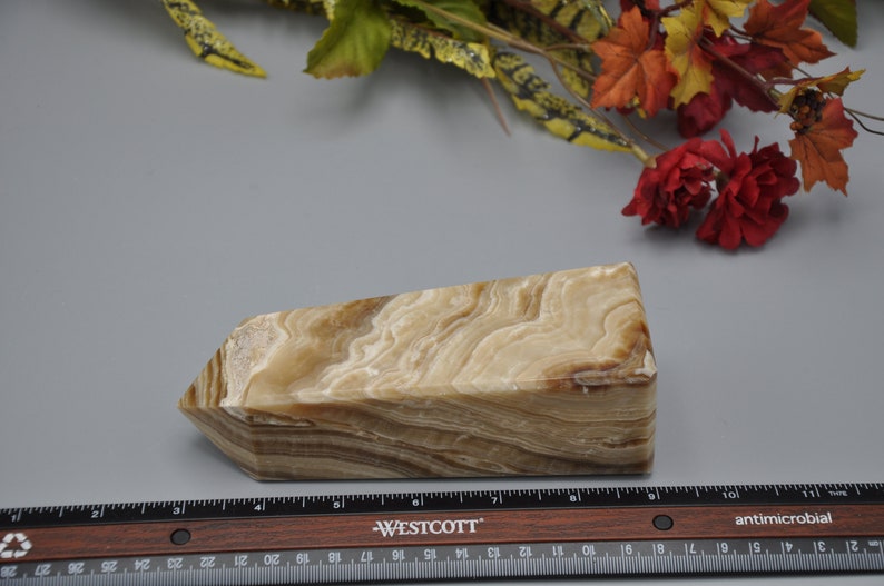May include: A rectangular, beige and brown onyx crystal with a layered pattern. The crystal is cut with angled sides and a flat top. A ruler with inch measurements is in the foreground.