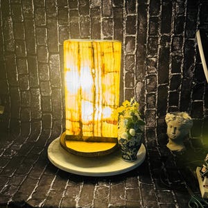 May include: A rectangular onyx lamp with a warm yellow glow, sitting on a wooden base and a white circular base. The lamp is in front of a black brick wall with a small bouquet of flowers and a white plaster statue of a woman's head.