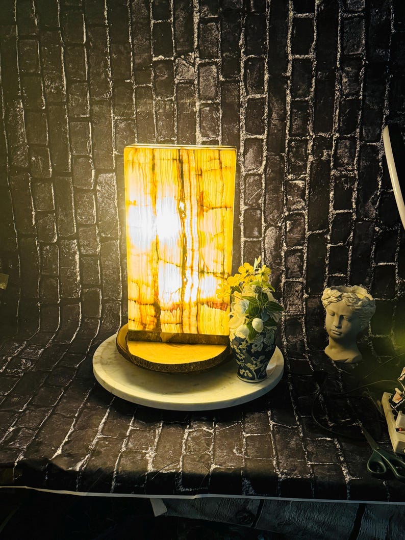 May include: A rectangular onyx lamp with a warm yellow glow, sitting on a wooden base and a white circular base. The lamp is in front of a black brick wall with a small bouquet of flowers and a white plaster statue of a woman's head.