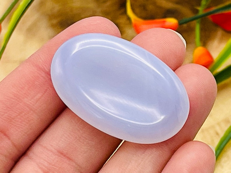 May include: A smooth, oval-shaped blue chalcedony gemstone. The stone is polished and has a light blue color.