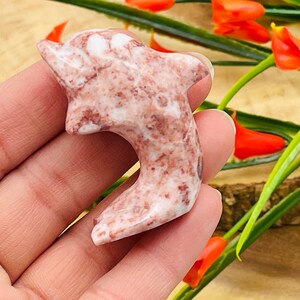 Puede incluir: Una mano sosteniendo una figurita de delf&iacute;n tallada en rosa y blanco. El delf&iacute;n est&aacute; hecho de una piedra marmoleada con un acabado liso y pulido.