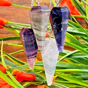 May include: Four crystal pendulums with silver chains. The pendulums are in various shades of purple, white, and green. The pendulums are hanging against a background of green leaves and orange flowers.