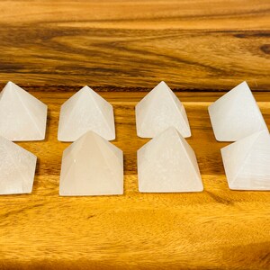 May include: Eight white selenite crystal pyramids arranged in two rows of four on a wooden surface.