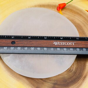 May include: A white selenite charging plate, approximately 7 inches in diameter, is shown on a wooden surface. A ruler is placed on top of the plate to show its size.