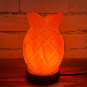 May include: A Himalayan salt lamp shaped like a pineapple with a warm orange glow. The lamp is on a wooden base and has a cord.