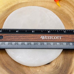 May include: A white selenite charging plate, approximately 6 inches in diameter, is shown on a wooden surface. A ruler is placed next to the plate for size reference.