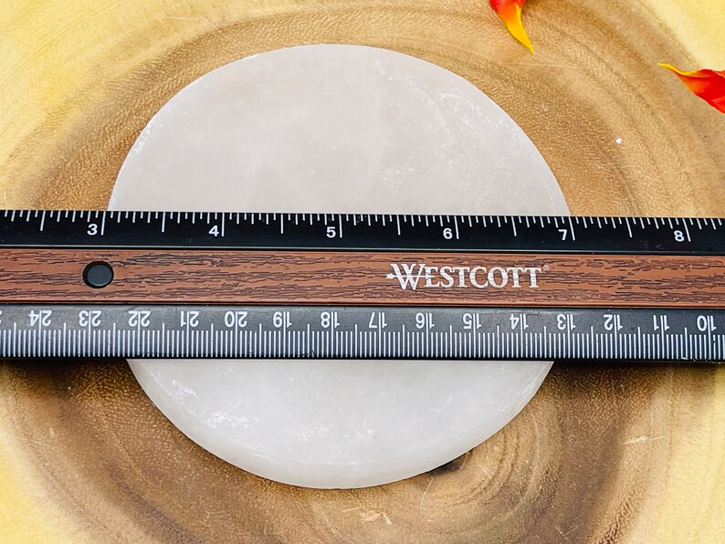 May include: A white selenite charging plate, approximately 6 inches in diameter, is shown on a wooden surface. A ruler is placed next to the plate for size reference.