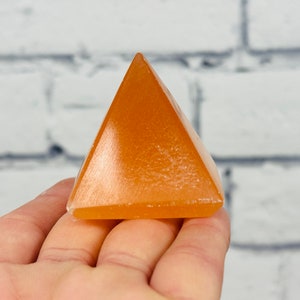 May include: A small, orange, crystal pyramid. The pyramid is smooth and polished, with a slight sheen.