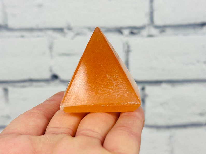 May include: A small, orange, crystal pyramid. The pyramid is smooth and polished, with a slight sheen.