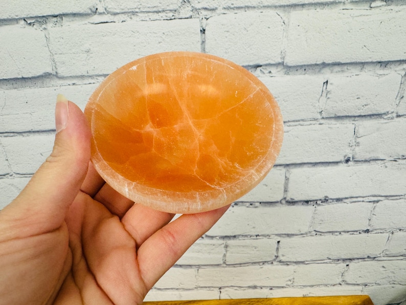 May include: A small, round, orange-coloured bowl made of selenite crystal. The bowl has a smooth, polished surface and a natural, organic shape.