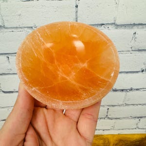 May include: A small, orange, crystal bowl with a smooth, polished surface. The bowl has a natural, organic shape and is perfect for holding small items like jewellery or crystals.