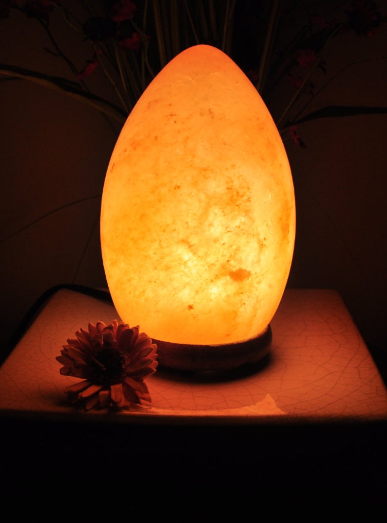 May include: A warm, orange glow emanates from a large, egg-shaped salt lamp. The lamp is made of natural Himalayan salt crystals and sits on a wooden base.