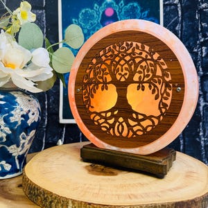 May include: A round Himalayan salt lamp with a wooden tree of life design. The lamp is lit and glows orange. The lamp is on a wooden base and is sitting on a wooden surface.